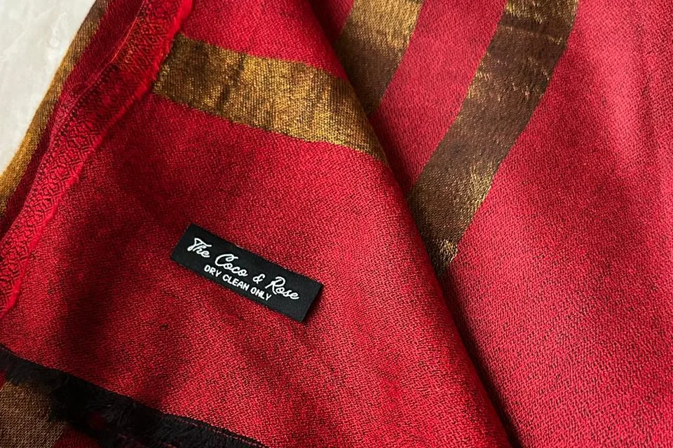 Una estola roja y dorada doblada, con su etiqueta tejida a la vista: The Coco & Rose, solo limpieza en seco